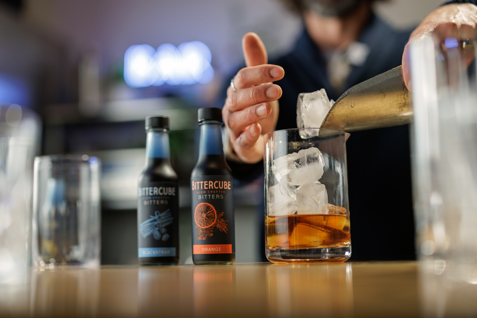 Ice being poured into a cocktail next to two bitters bottles